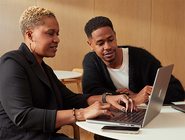 Dos colegas de Apple sentados frente a una mesa, colaboran usando una MacBook. 