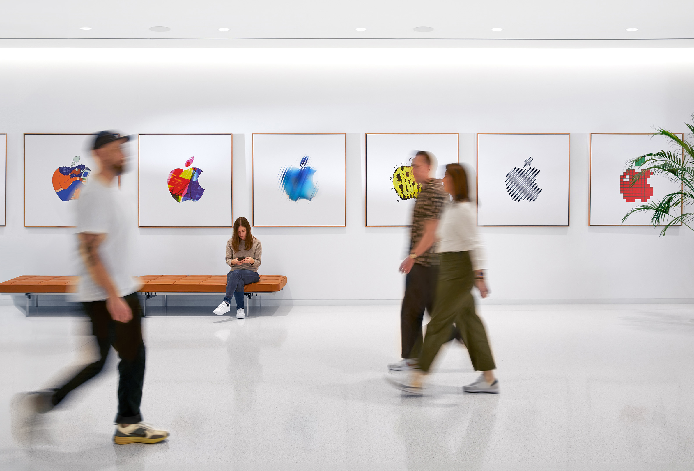 Tres colegas de Apple pasan caminando frente a una pared interior con logotipos coloridos de Apple, una cuarta integrante del equipo está sentada en una banca.