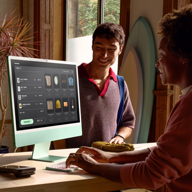 Una persona, sentada detrás del mostrador de una tienda que vende tablas de surf y otros artículos, usa un iMac para completar la compra de un cliente que está del otro lado del mostrador.